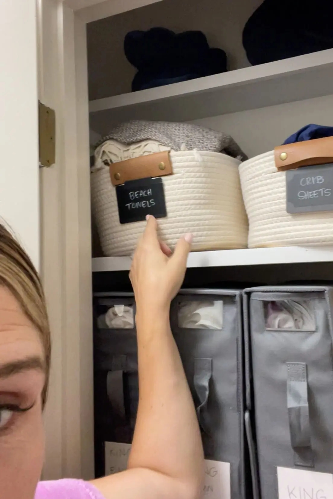 Close-up of a hand pointing to a labeled woven basket for beach towels in an organized linen closet.